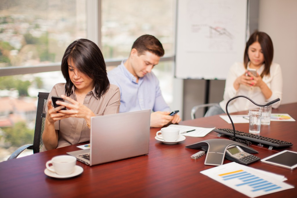 Hispanic people in a meeting room ignoring their work and doing some social networking on their smartphones